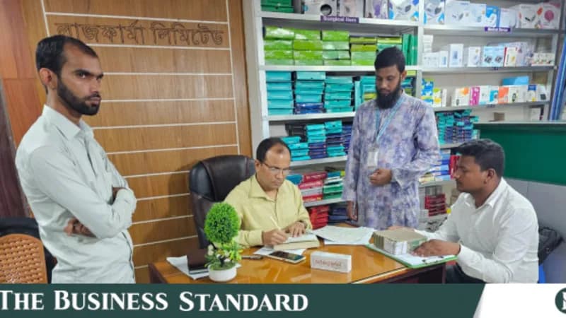 The interior of a retail pharmacy in Bangladesh, staff behind a counter lined with shelves of boxed medicine.